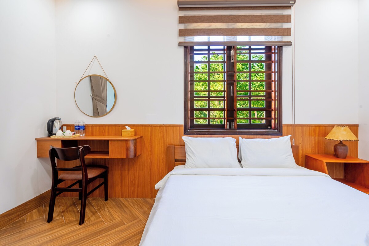 Une chambre d’hôtel comprenant un lit, un bureau avec une chaise, un miroir, une fenêtre donnant sur un espace verdoyant et une table de chevet avec une lampe.