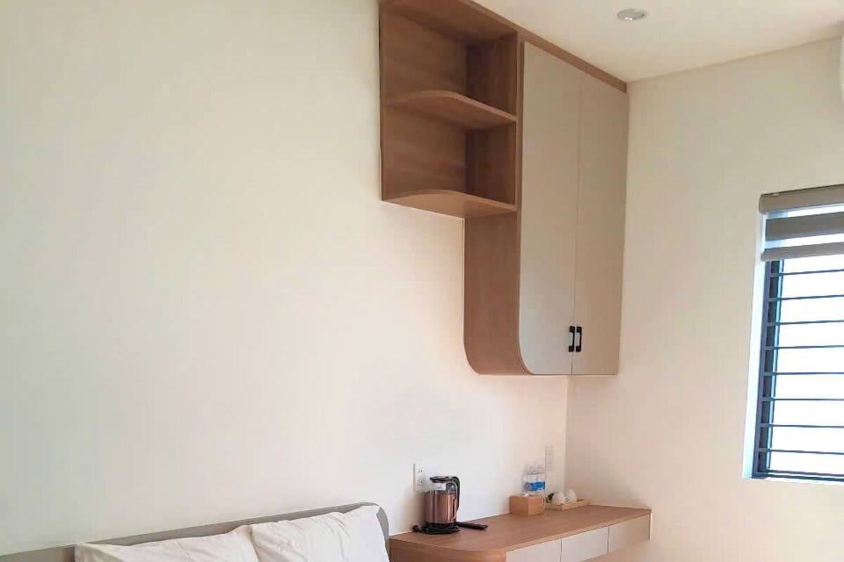 Une chambre avec un lit, une étagère intégrée, une petite table avec une bouilloire et une fenêtre avec des stores.