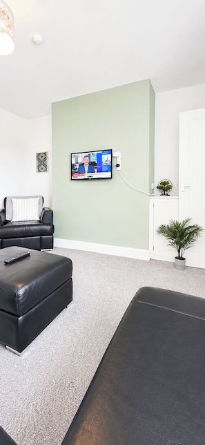 A living room with a TV, a sofa, an ottoman, a small plant, and a door. - House Sleeps 7 with WiFi (Gateshead)