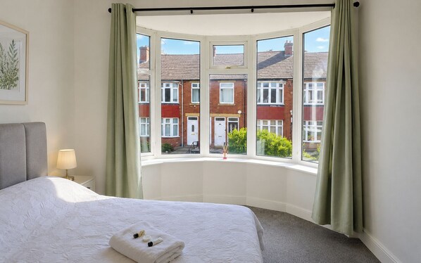 A bedroom with a large bed, a bedside table with a lamp, a window with curtains, and a view of red brick buildings. - House Sleeps 7 with WiFi (Gateshead)