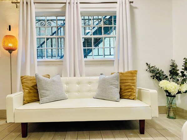 A white sofa with two grey and two gold cushions, a floor lamp, and a vase with white flowers.