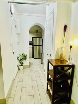 A narrow hallway with white walls, a wooden table with a vase and candle, and a kitchen in the background.