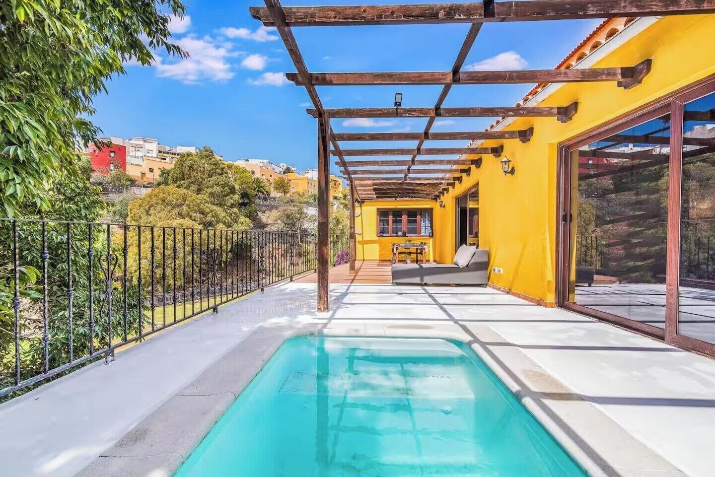 Une piscine avec une pergola, un bâtiment jaune et une vue sur les environs.