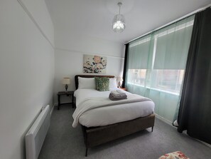 Ein Schlafzimmer mit Bett, Nachttischen, einer Hängelampe, einem Fenster mit Vorhängen und einem Wandbild.