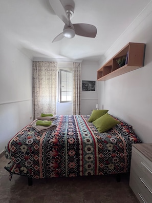 Ein Schlafzimmer mit einem Bett, einem Deckenventilator, einem Fenster mit Vorhängen, einem Wandregal und einer Tür zu einem weiteren Raum.
