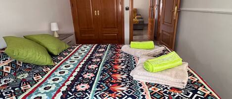 Une chambre à coucher avec une couvre-lit à motifs, un coussin vert, une serviette pliée et une armoire en bois.