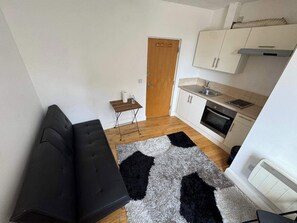 Ett kompakt bostadsutrymme med en soffa, ett litet bord, en köksdel med skåp och vitvaror samt en dörr.