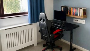 Quarto com escrivaninha, computador, cadeira, radiador, estante de livros e janela com cortinas.