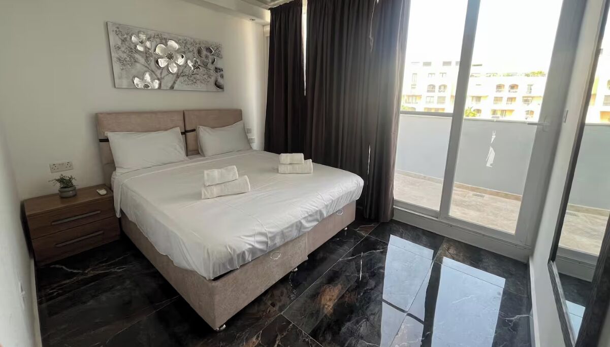 A bedroom with a bed, a nightstand, a wall art piece, and a view of a building through a window.