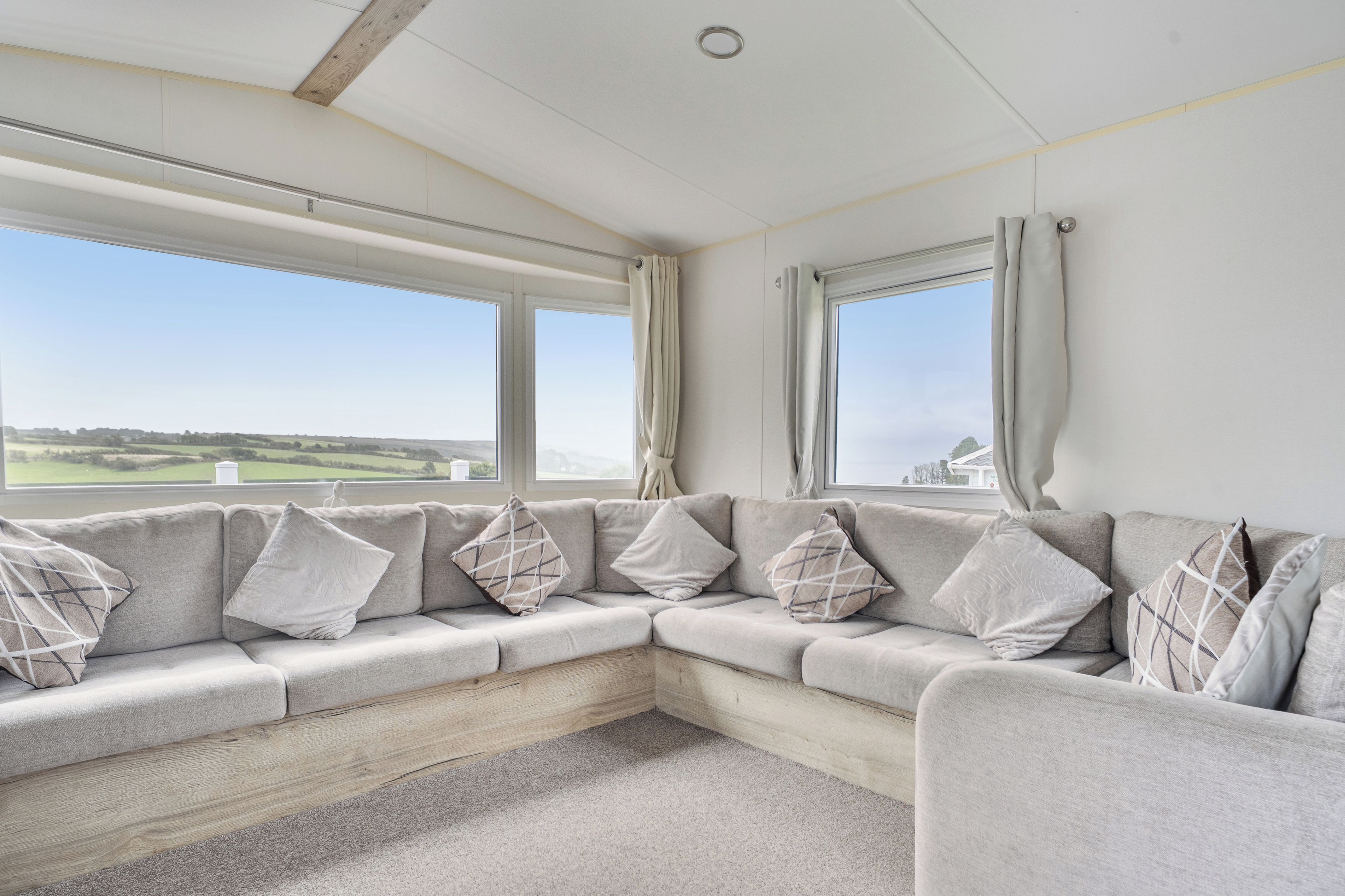 Ein modernes Wohnzimmer mit einem großen, winkelförmigen Modulsystem, zwei Fenstern mit Vorhängen und Blick auf die Landschaft.