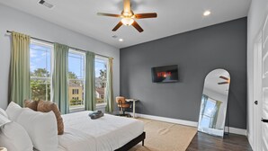 Une chambre à coucher avec un lit, un ventilateur de plafond, une télévision, une chaise et une grande fenêtre avec des rideaux.