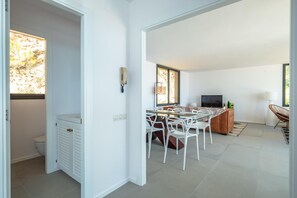 A modern kitchen with white cabinetry, a dining area with white chairs, a TV, and a window with a view of trees.