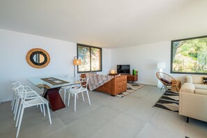 A modern living room with a glass dining table, white chairs, a brown sofa, a TV, and large windows.
