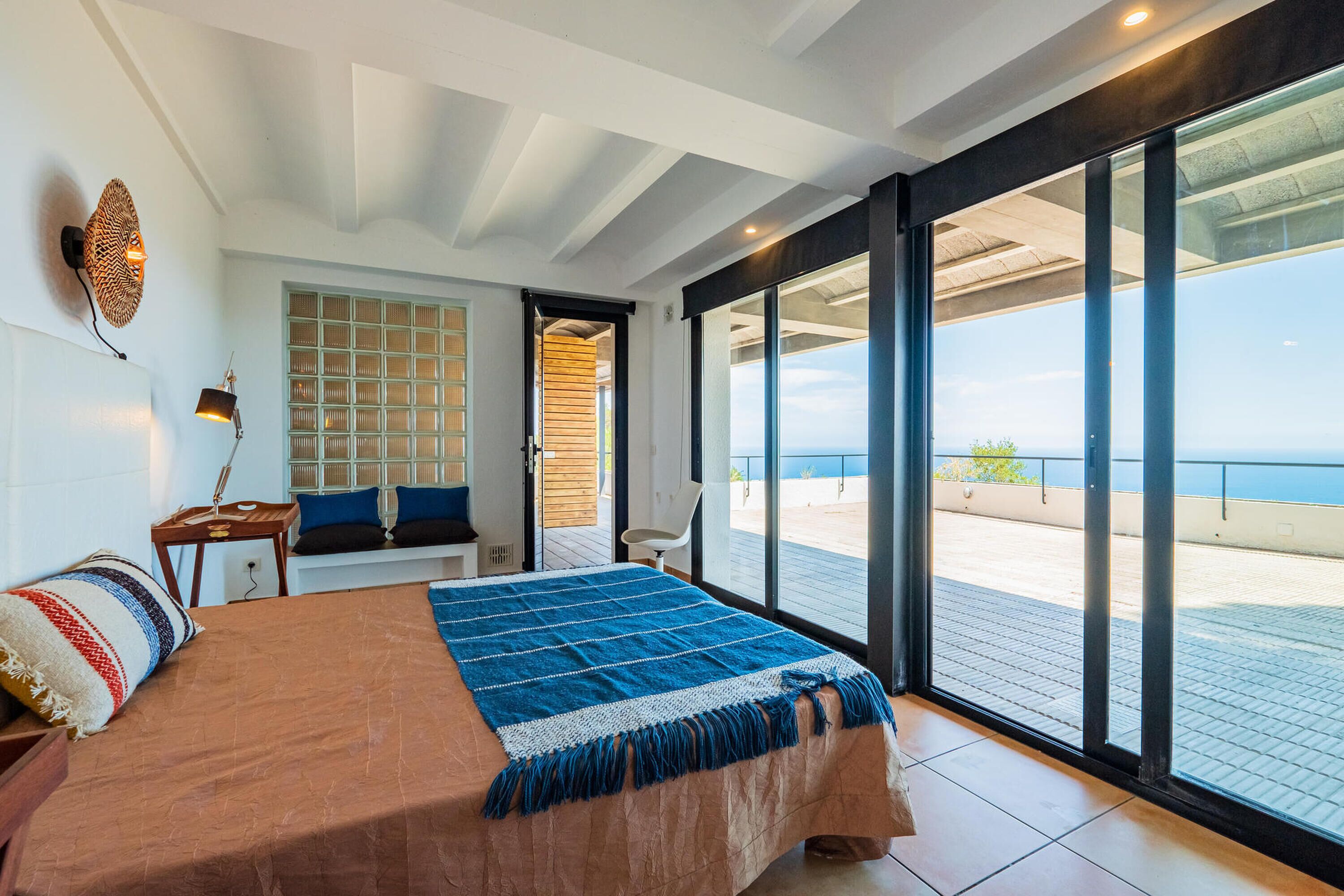 A bedroom with a large bed, a bench, a wall-mounted lamp, and a view of the sea through large sliding doors.