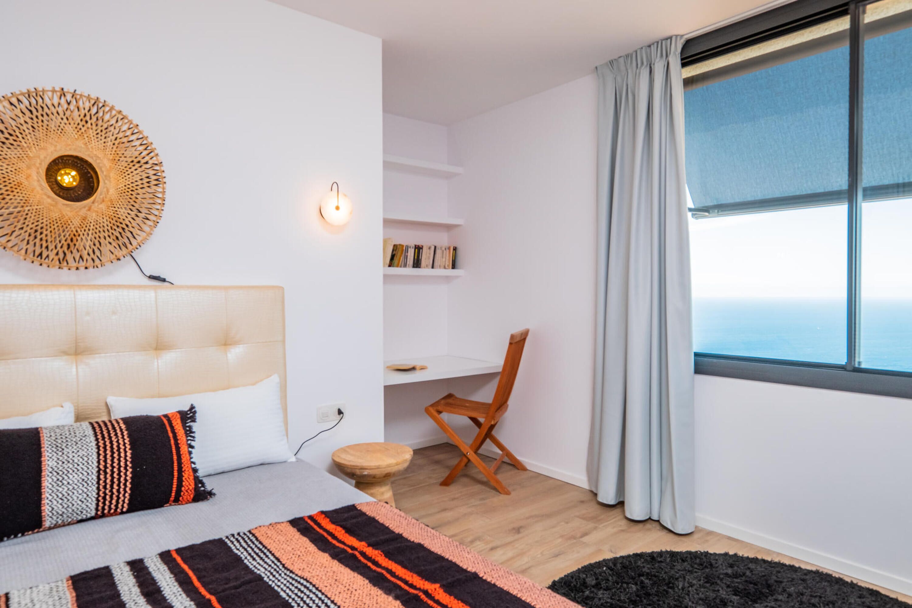 A bedroom with a bed, a desk, a chair, a round wall decoration, and a large window.