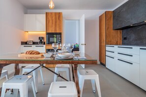 A modern kitchen with white cabinets, a wooden accent wall, a glass dining table, and white stools.