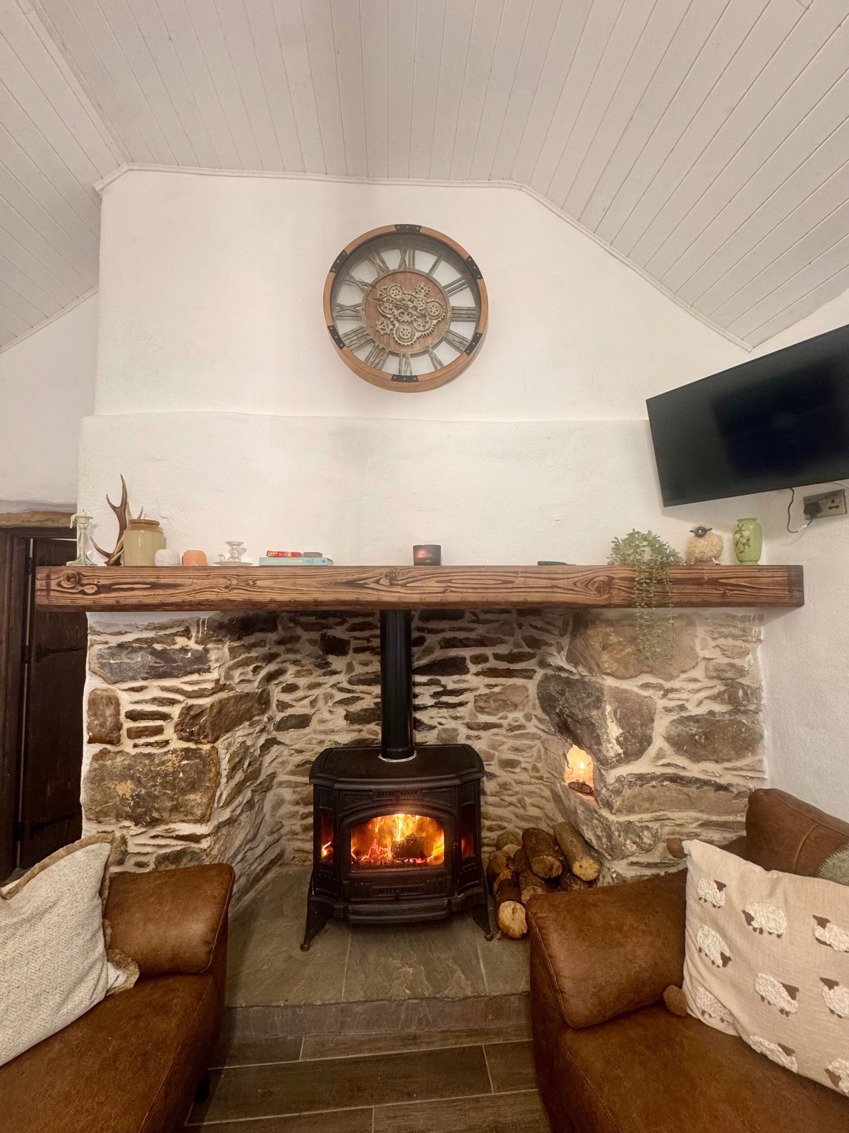 Ein Kamin aus Stein mit einer modernen Uhr darüber, ein Flachbildfernseher an der Wand und eine gemütliche Sitzecke mit braunen Kissen.