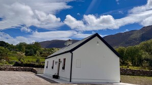 A white building with a gabled roof, a stone wall, and a grassy landscape with mountains in the background.