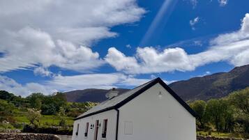 A white building with a gabled roof, a stone wall, and a grassy landscape with mountains in the background.