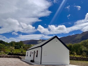 A white building with a gabled roof, a stone wall, and a grassy landscape with mountains in the background. - Cottage Sleeps 4 with Parking (Killarney)