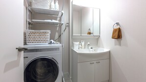A compact laundry area with a washing machine, shelves with laundry baskets, a sink, and a mirror. - Kasatori-cho I [9-minute drive from Nagoya Station] Newly Built 1LDK / WiFi Included / Full Amenities / Sleeps Up to 3 / Ideal for Sightseeing / 1-101 (Kasatori-cho, Nishi-ku, Nagoya City,)