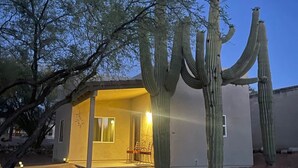 A desert landscape with a large saguaro cactus in the foreground and a modern house with a patio in the background. - Saguaro Grove, Built May '26, Smart Lock, Quiet Cul-de-sac (Tucson)
