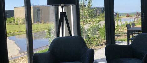 A room with large windows, a black floor lamp, two dark armchairs, and a view of a building and greenery outside.