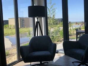 Een kamer met grote ramen, een zwarte vloerlamp, twee donkere fauteuils en uitzicht op een gebouw en groen.