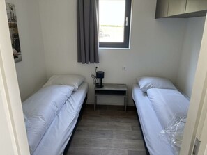 Een kleine, schone kamer met twee enkele bedden, een tafeltje met een lamp en een raam met gordijnen.