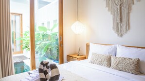 Een hotelkamer met een bed, een nachtkastje, een lamp, een wanddecoratie van macramé en een schuifdeur naar een balkon met planten.