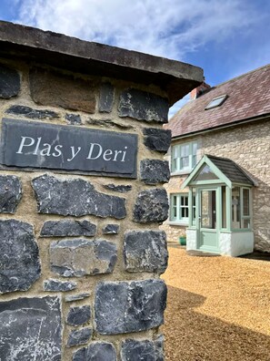 Eine Steinsäule mit dem Schild 'Plas y Deri' und ein traditionelles Steinhaus mit einem grün verglasten Anbau.