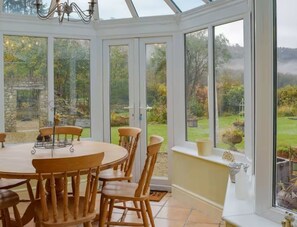 Ein Wintergarten mit rundem Tisch und Stühlen, großen Fenstern und Blick auf einen Garten.