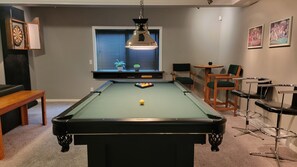 Una mesa de billar con una bola amarilla, rodeada de sillas y una mesita, con una ventana y persianas al fondo.