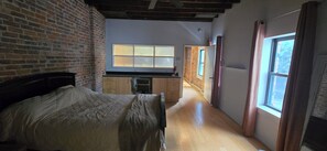 A bedroom with a bed, a brick wall, a window, and a kitchen area with cabinets and a countertop.
