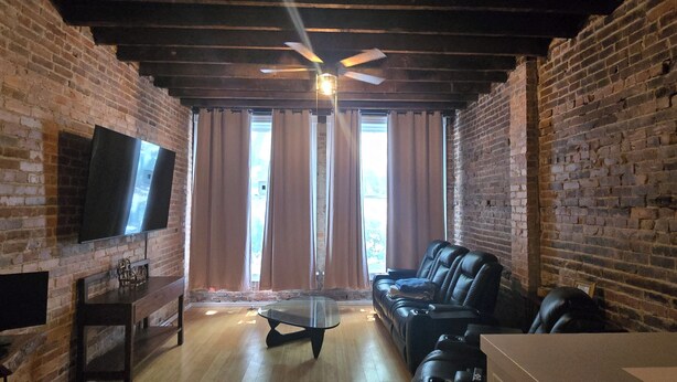 Ein Zimmer mit Ziegelwänden, einem Deckenventilator, einem Flachbildfernseher, einem Ledersofa und einem Holztisch.