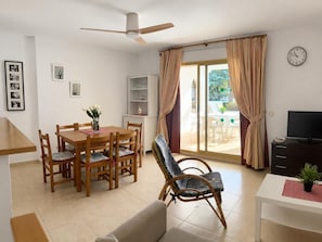 Ein Wohnzimmer mit Esstisch, Stühlen, einer Couch, einem Fernseher, einem Bücherregal und einer Glasschiebetür, die zu einem Außenbereich führt.