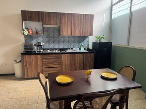 A compact kitchen with wooden cabinets, a microwave, a coffee maker, and a refrigerator. There's a dining table with two yellow plates and a small decorative bird.