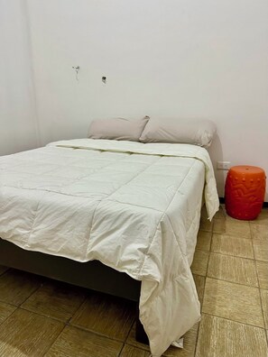 A single bed with a white bedspread and pillows, a red stool, and a tiled floor.