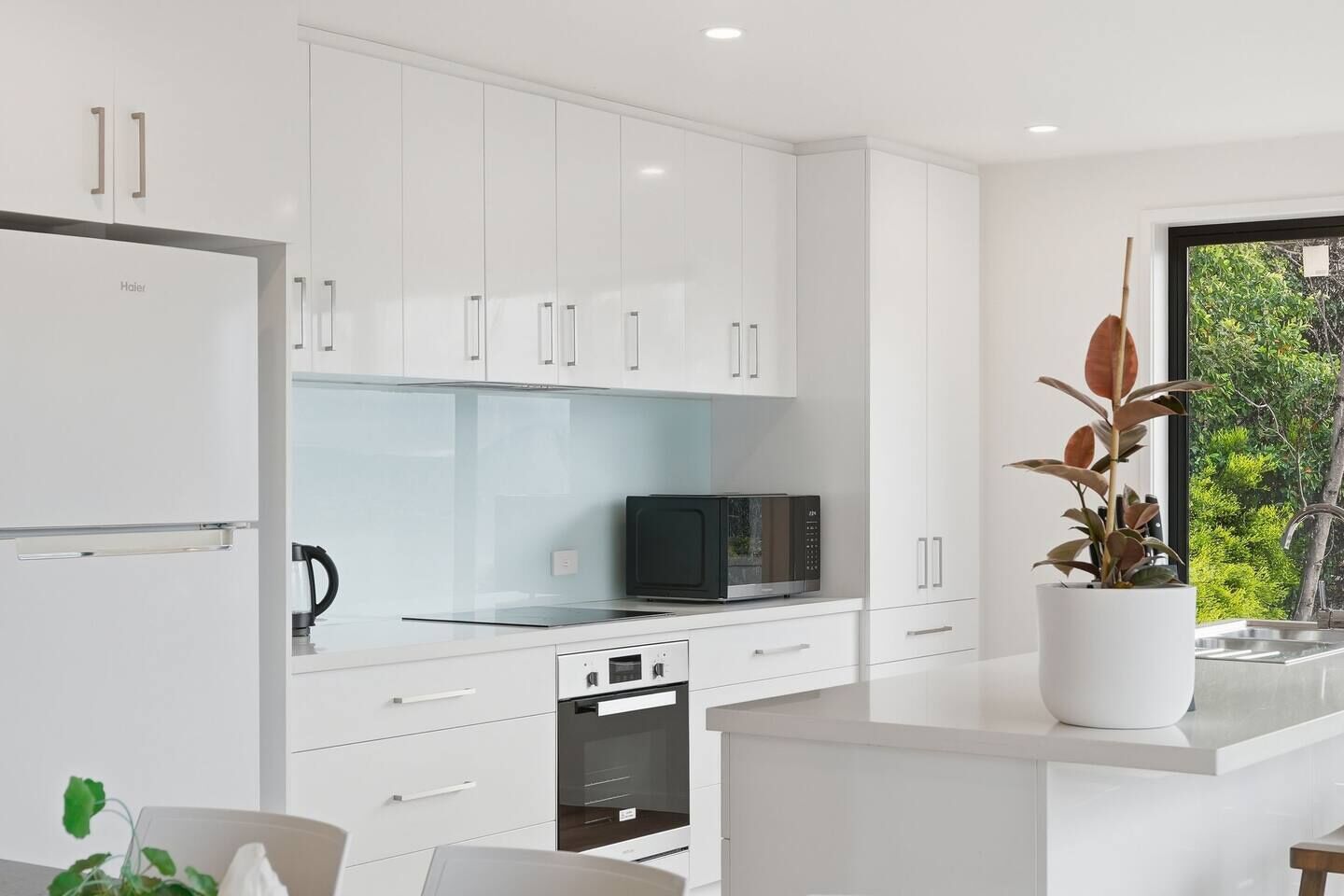 A modern kitchen with white cabinetry, a microwave, a kettle, and a potted plant.