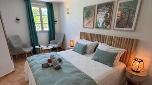 A bedroom with a large bed, two bedside tables, a chair, a small table, and a window with curtains. - LARIMAR APARTMENT (Saint Martin)