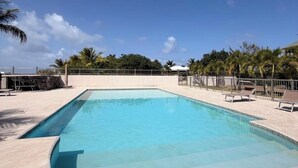 A rectangular swimming pool with a tiled deck, surrounded by palm trees and a clear sky. - LARIMAR APARTMENT (Saint Martin)