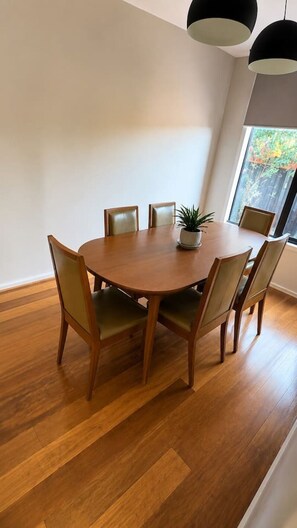 Een ronde houten eettafel met zes stoelen, een potplant en twee hanglampen.