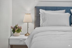 Una cama bien hecha con ropa de cama blanca, un cabecero azul y una mesita de noche con una lámpara y una planta en maceta.