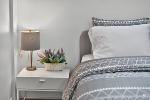 A neatly arranged bedroom with a bedside table, a lamp, a potted plant, and a bed with patterned bedding. - Modern and spacious 3-BR basement suite (Vancouver)