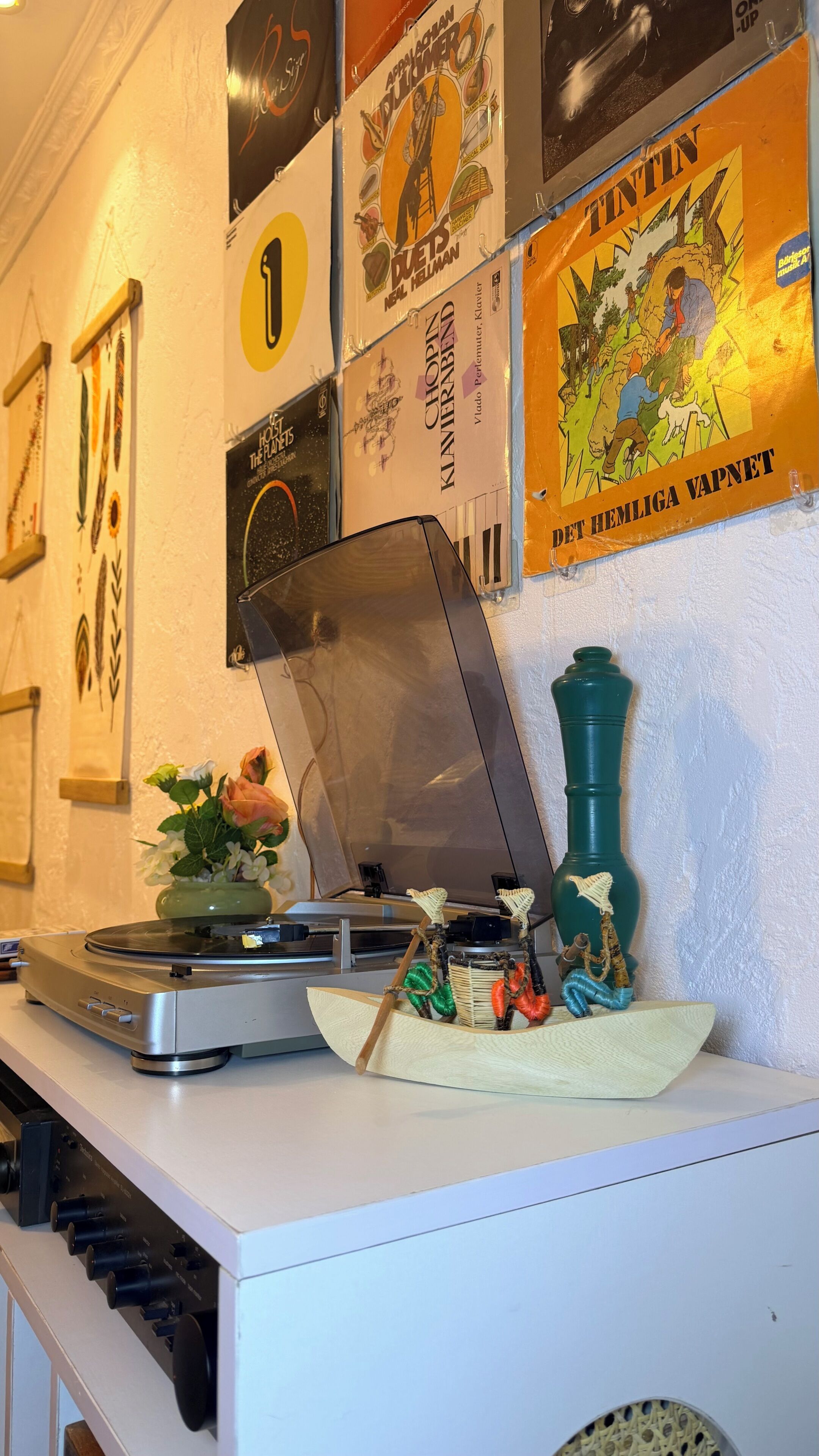 Un tocadiscos con discos de vinilo, un barco decorativo y un jarrón verde sobre una mesa blanca.