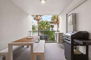 Ein Balkon mit einem Holztisch und Bänken, einem Grill sowie Blick auf Palmen und Gebäude.