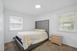 Une chambre à coucher avec un lit, une commode et deux fenêtres équipées de stores.