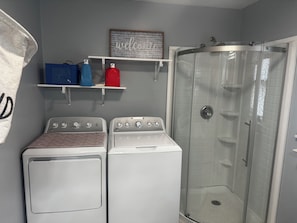 Un espace buanderie comprenant deux machines à laver à chargement frontal, un espace douche et des étagÚres de rangement avec divers articles.
