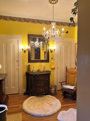 Une piĂšce avec un lustre, un miroir, une coiffeuse avec un lavabo, une chaise et un tapis rond.