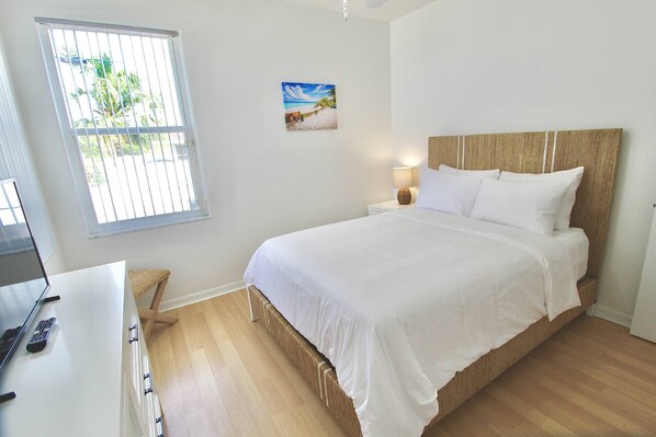 Un dormitorio con cama, escritorio con computadora portátil, silla, ventana con rejas e imagen colgada en la pared.